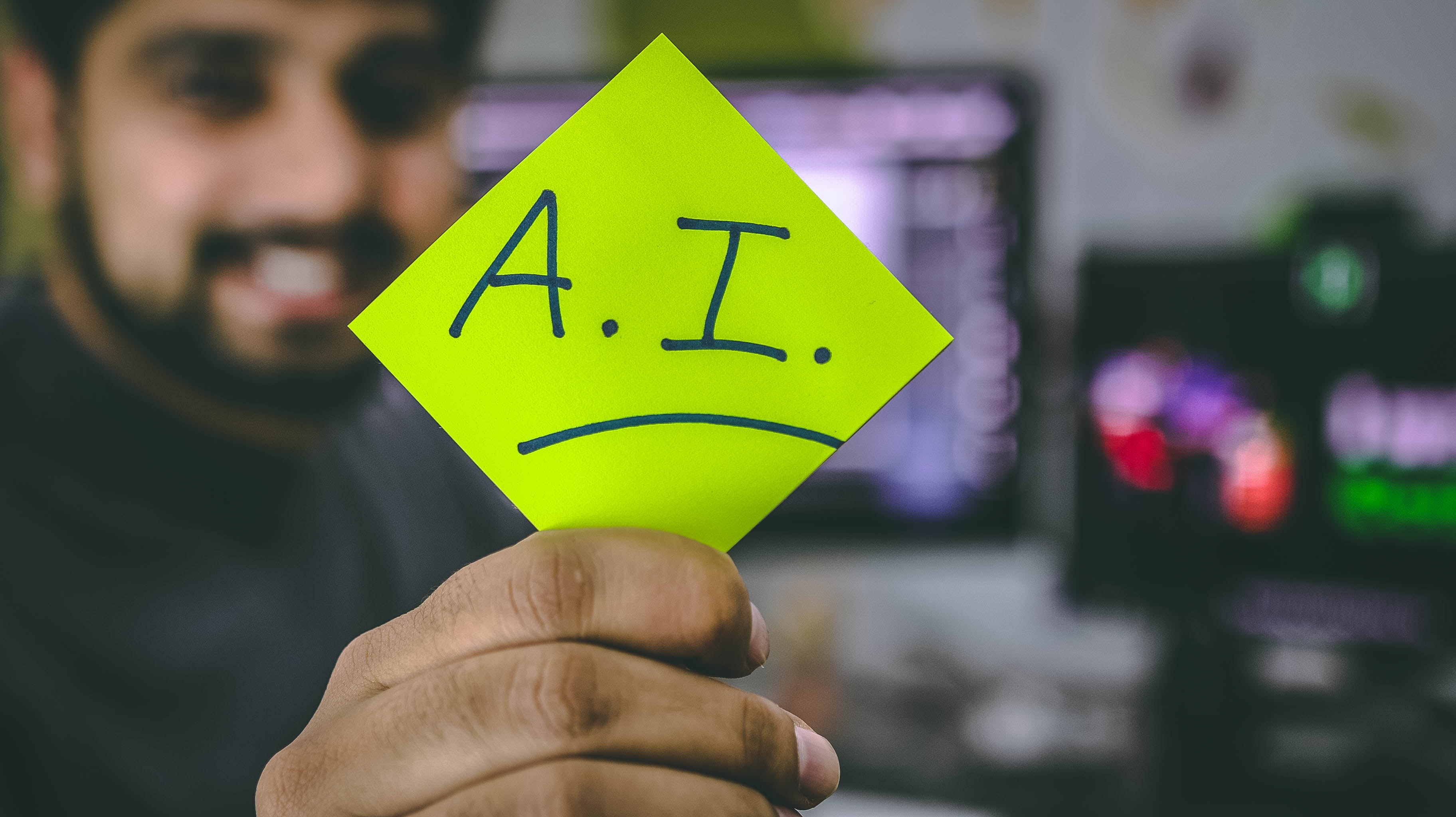Photograph of a computer programmer holding up a sticky note that says "A.I."