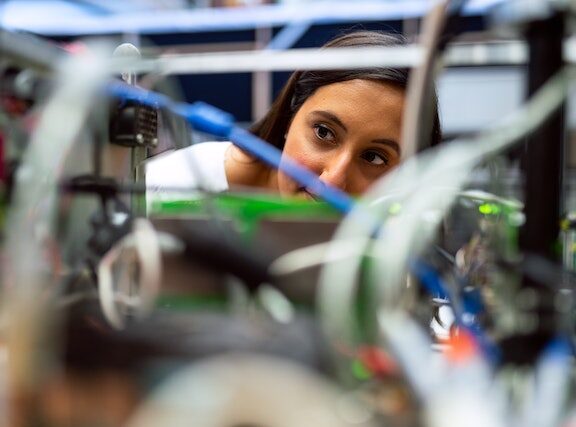 A photograph of a female engineer peering through the wires of a computer.