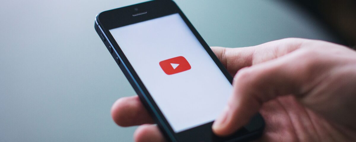 Photograph of a hand holding a smartphone. The smartphone displays the Youtube logo.