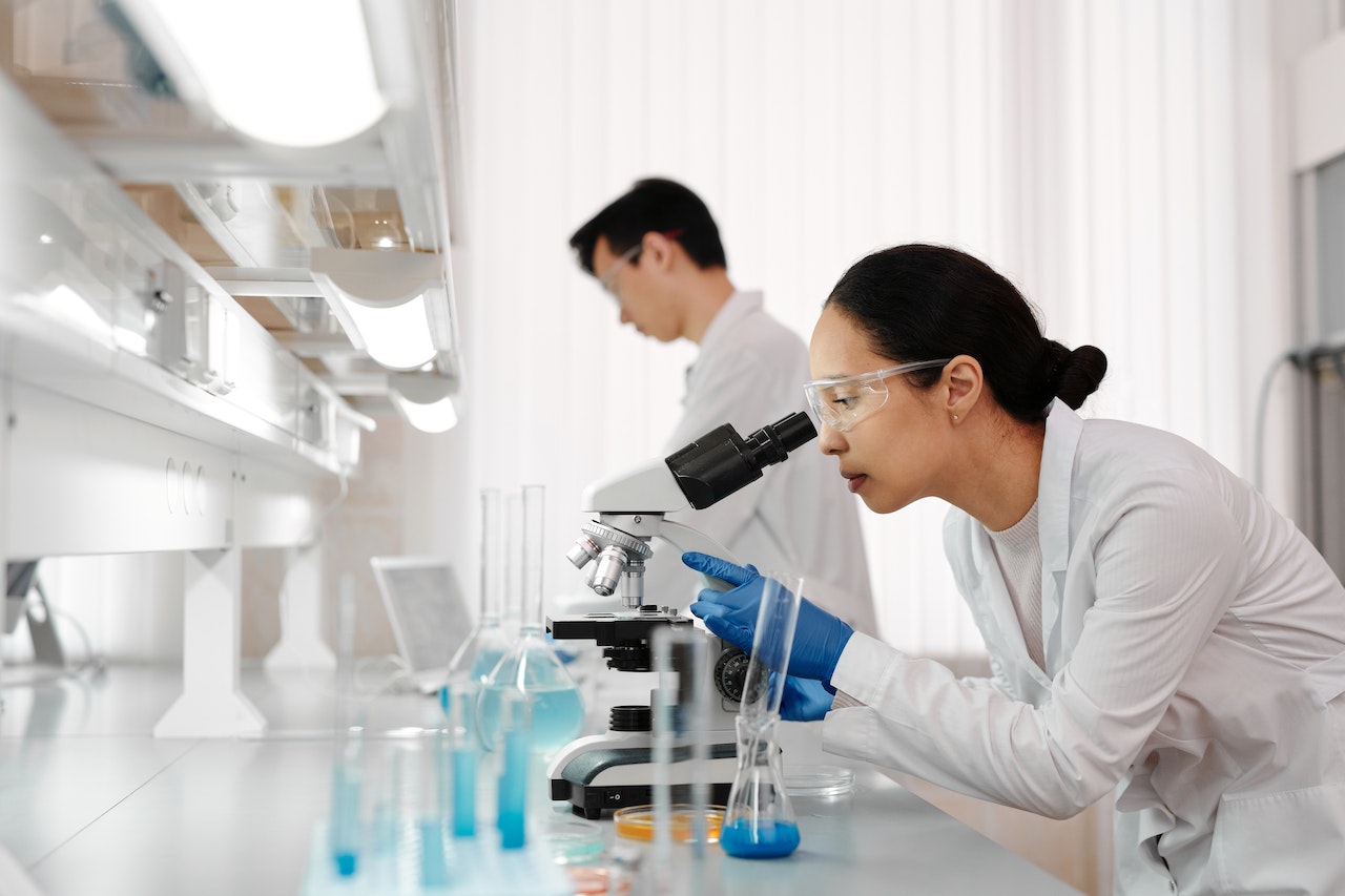 Photograph of two inventors wearing lab coats in a scientific laboratory. The nearest inventor is looking into a microscope. There are beakers on the counter beside her.