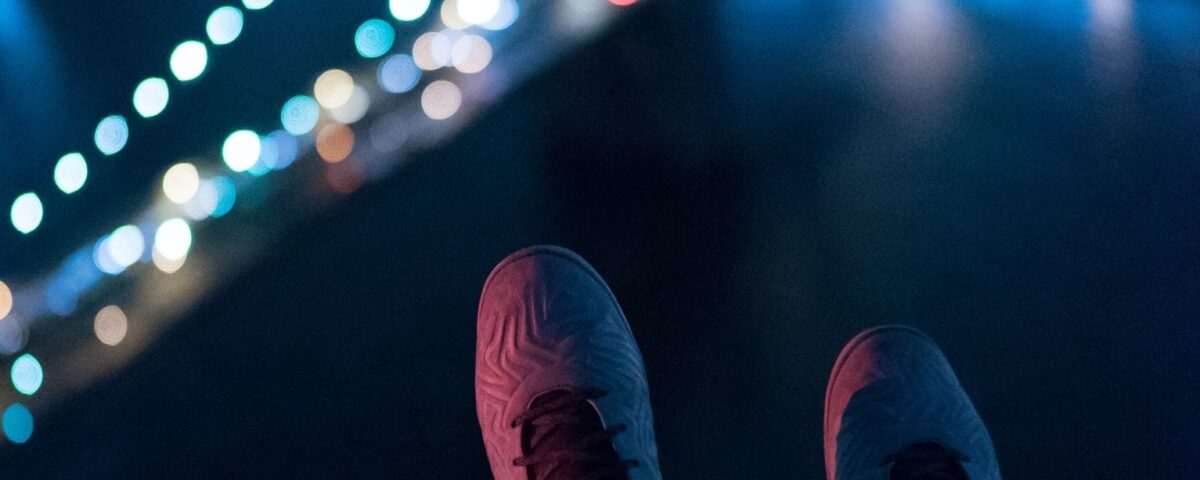 Photo of a pair of shoes dangling off the edge of a building while lights shine on a bridge in the distance at night.