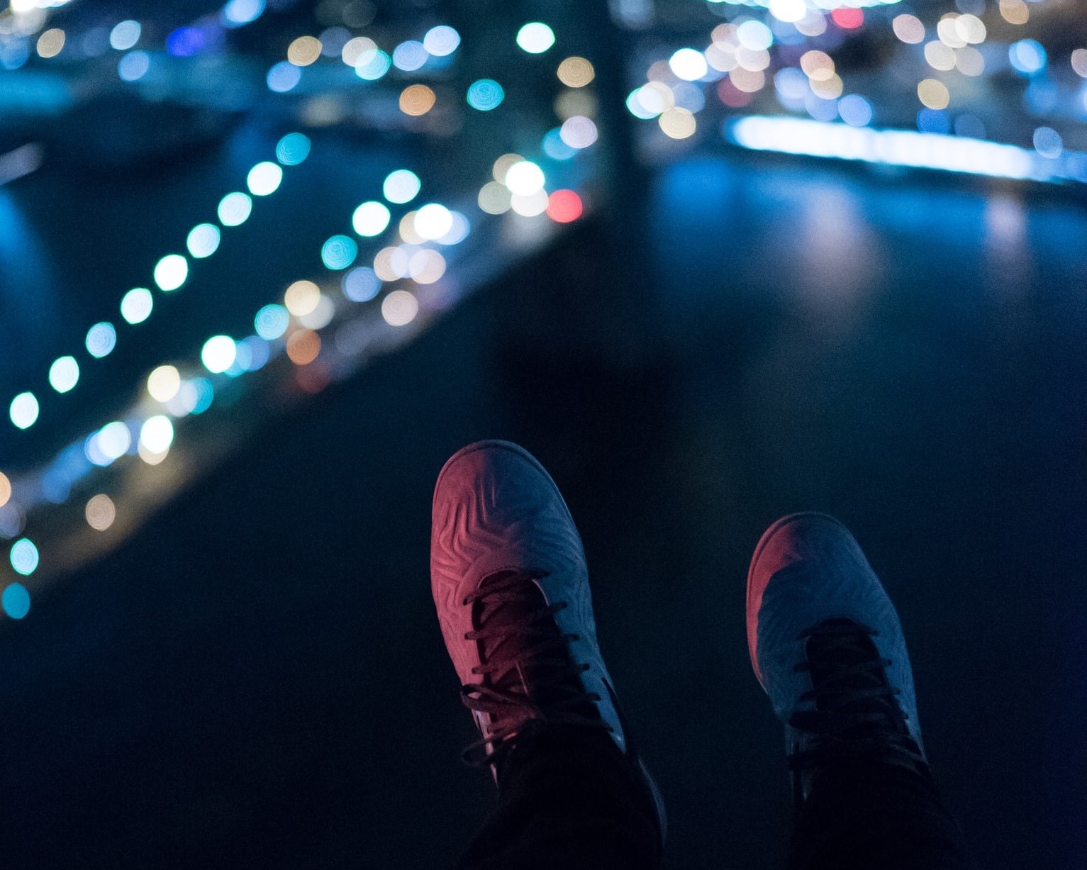 Photo of a pair of shoes dangling off the edge of a building while lights shine on a bridge in the distance at night.