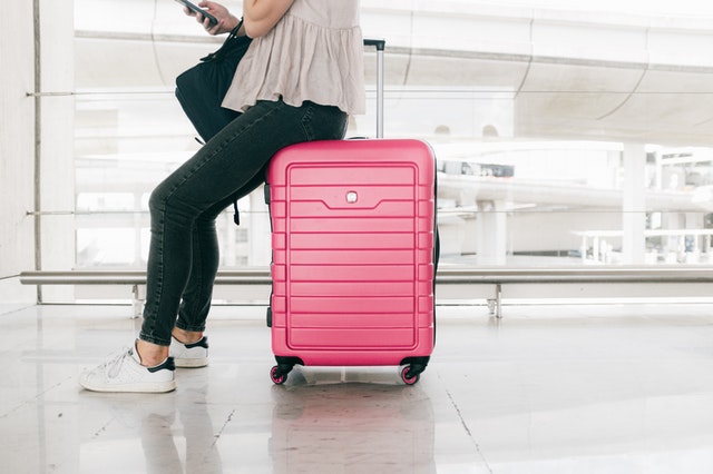 A woman sits on a pink suitcase in an airport.