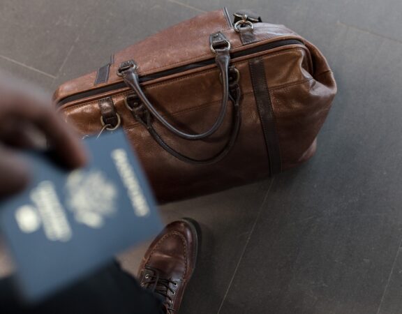 Photograph of a man holding a passport with leather luggage on the ground at his feet.