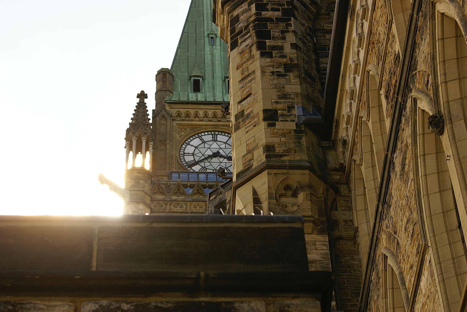 Photograph of the Peace Tower at the Canadian parliament buildings taken from the ground so that the clock peeks out from behind another building.