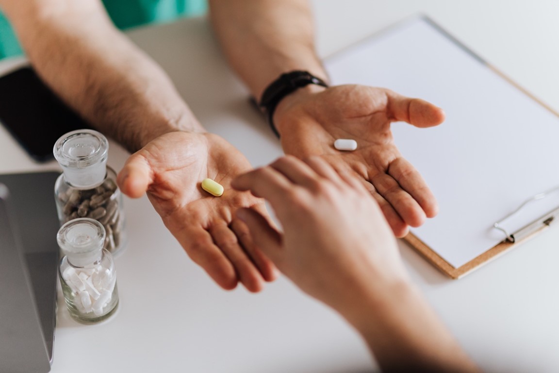 Photo of a pharmacist holding out a yellow pill in one hand and a white pill in another hand. A patient reaches for the yellow pill.